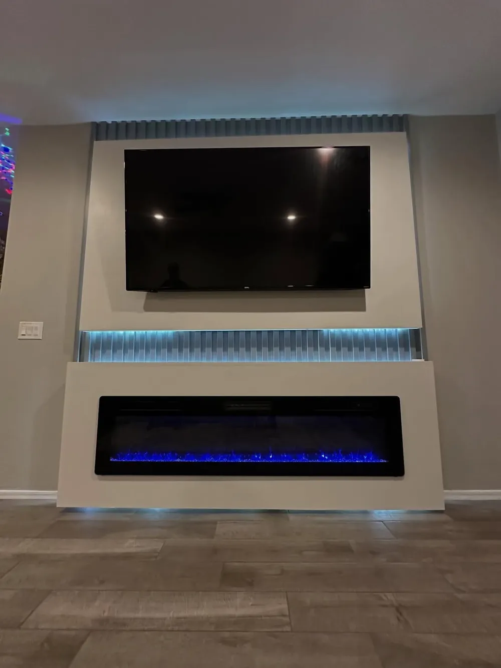 A fluted panel wall in soft greige, floating television above a blue lit linear fireplace.